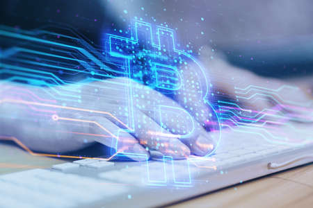 Close up of hands using computer keyboard with creative blue bitcoin hologram on blurry office interior background. Cryptocurrency and technology concept. double exposureの写真素材