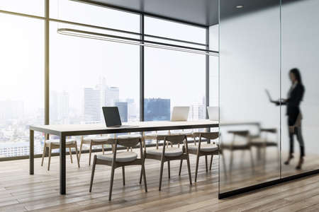 Attractive young european businesswoman with laptop standing in modern concrete office interior with wooden flooring, matte partition glass, furniture, equipment and city view with daylight. Worker and ceo conceptの写真素材