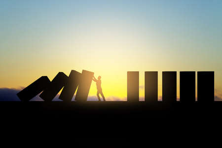 Backlit business man stopping abstract wooden domino blocks on bright sunset sky background with mock up place. Crisis, recession and challenge conceptの写真素材