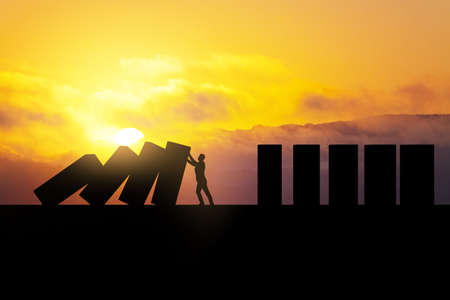 Backlit man stopping abstract wooden domino blocks on bright sunset sky background with mock up place. Crisis, recession and challenge conceptの写真素材