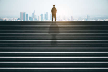 Back view of businessman on top of concrete stairs looking into the distance on daylight city background. Success and future conceptの写真素材