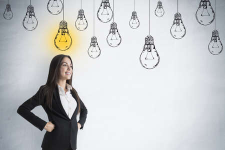 Portrait of attractive pensive young european businesswoman standing on concrete wall background with glowing drawn light bulb sketch and mock up place. idea and creativity conceptの写真素材