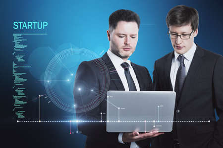 Attractive young european businessmen using laptop with futuristic blue HUD interface and glowing programming code on blurry blue background. Digital start up, teamwork, future and technology conceptの写真素材