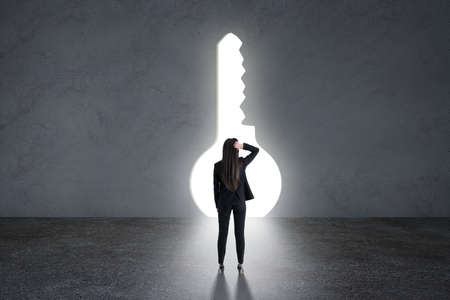 Find a solution and way concept with back of pensive businesswoman grabbing her head in black suit looking at big bright and glowing keyhole in gray wallの写真素材