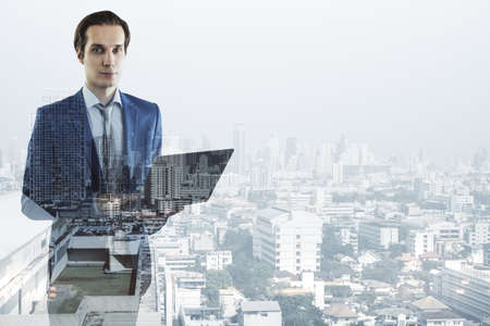 Thoughtful young businessman holding laptop on blurry city background with mock up place. Success, think, future and ceo concept. double exposureの写真素材