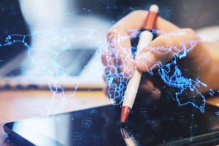 Close up of hands using digital tablet with pen and laptop with glowing blue map hologram and...