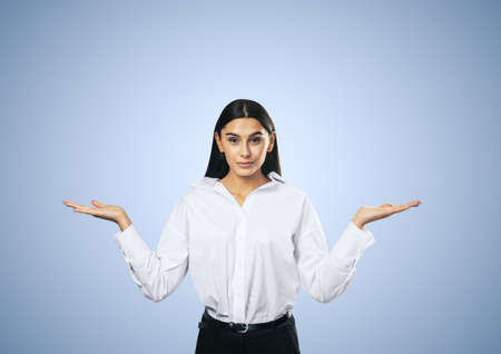 Right choice concept with front view on confused young woman in white shirt isolated on abstract light blue wall background, closeupの写真素材