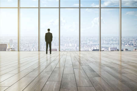 Business success, leadership and inspiration concept with businessman back view looking on city skyline through big window in empty spacious hall with wooden floorの写真素材