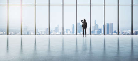 Thinking about future and career opportunities concept with pensive businesswoman back view looking through big window on sunny city in empty space with glossy concrete floorの写真素材