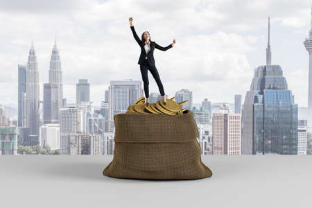 Attractive young european businesswoman celebrating success on top of huge sack filled with golden coins on light city background. Wealth, treasure and boss conceptの写真素材