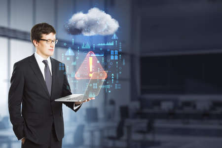 Attractive young european businessman with laptop standing in blurry office interior with abstract hologram raining cloud and mock up place. Data, information leak, security and theft concept. double exposureの写真素材