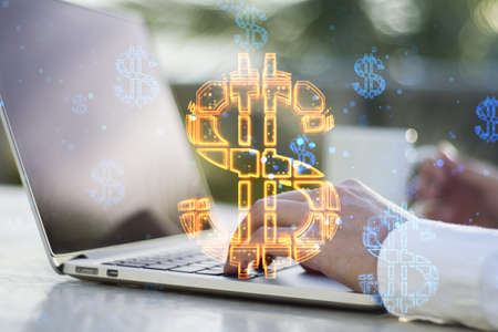 Close up of hands using laptop keyboard with abstract glowing dollar sign hologram on blurry desktop...