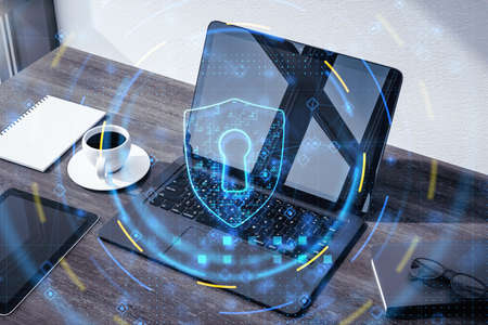 Data safety and cyber security concept with digital graphic glowing blue shield symbol with keyhole on office table with laptop and coffee mug background, double exposureの写真素材
