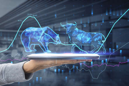 Stock exchange trading concept. The bulls and bears struggle. Equity market illustration. Close up of hand holding tablet with creative hologram and graph on blurry office interior background. double exposureの写真素材