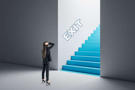Thoughtful young businesswoman in concrete interior looking at blue stairs exit. Way out, solution and choice conceptの写真素材