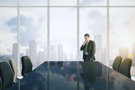 Attractive young european businessman thinking in modern meeting room interior with reflections on table, chairs and panoramic window with sky clouds view and daylight. CEO and conference conceptの写真素材