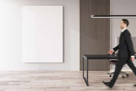Side view of young european businessman walking in office interior with furniture, equipment, wooden flooring and empty white mock up poster on concrete wall. CEO, executive and worker conceptの写真素材