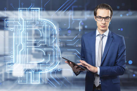Attractive young european businessman with glasses using tablet with creative glowing bitcoin...