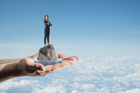 Abstract image of hands holding female standing on rock and water on bright blue sky city background with clouds and mock up place. Success, challenge and abstract conceptの写真素材