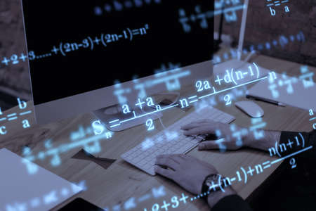 Close up of businessman hands using computer keyboard at workplace with monitor, supplies and glowing mathematical formulas on blue background. Education, knowledge and statistics conceptの写真素材