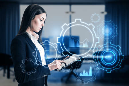 Attractive young european businesswoman with tablet and glowing cogwheel and chart hologram on blurry office interior background. Business analytics and finance concept. double exposureの写真素材