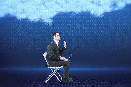 Attractive young european businessman with laptop sitting on blurry digital blue background with abstract cloud. Cloud computing, big data and digital transformation conceptの写真素材