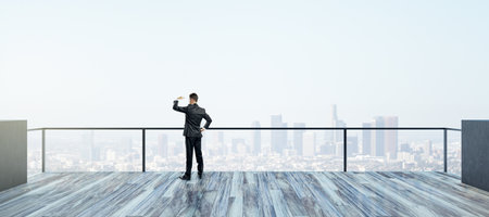 Back view of young businessman looking into the distance on rooftop with bright daylight sky and wide city view. Success, future and vision conceptの写真素材
