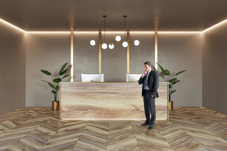 Front view on pensive businessman near stylish reception desk made from natural wooden slab with modern computers on parquet floor with green plants on beige wall background with lightning on topの写真素材
