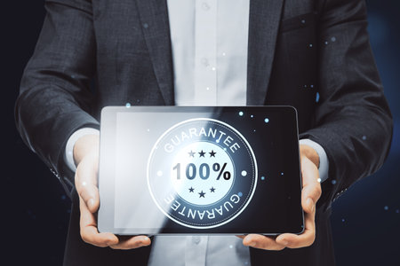 Close up of businessman hand holding tablet with glowing 100% guarantee stamp hologram on dark background with bokeh circles. Quality seal and warranty conceptの写真素材