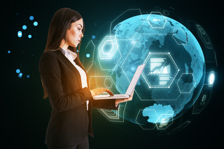 Side view of young businesswoman using laptop with digital business globe hologram on blurry background. Global technology, finance and web safety conceptの写真素材