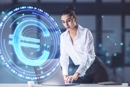 Attractive young businesswoman using laptop while leaning on desk with abstract blue digital euro coin on blurry office background. Online banking and trade concept. double exposureの写真素材