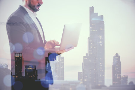 Young European businessman using laptop on blurry city background. Success, future and CEO concept. Double exposureの写真素材