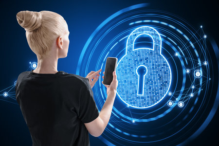 Back view of young blonde woman using cellphone with glowing polygonal padlock hologram on blurry dark background. Secure, safety and technology conceptの写真素材