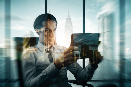 Attractive young European businessman silhouette sitting at desk and using tablet on abstract blurry city background. Success, workplace, CEO and future concept. Double exposureの写真素材