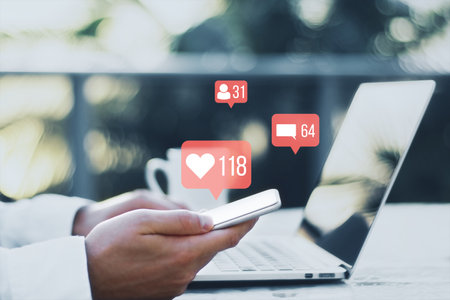 Close up of businessman hands using laptop and phone at desk with coffee cup with creative social media icons hologram on blurry outdoor background. Community, likes, shares and communication conceptの写真素材