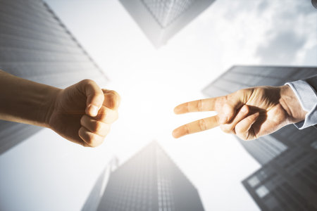 Close up of young businessmen hands playing rock paper scissors on bright city buildings background....