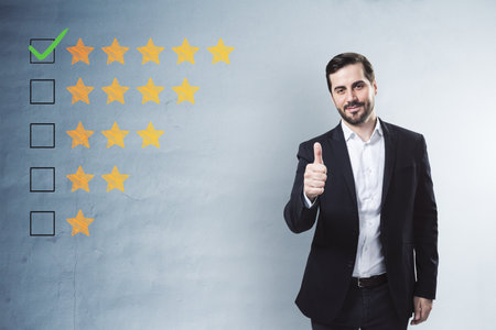 Happy young businessman showing thumbs up to 5 star rating on concrete wall background. Customer service and excellent feedbackの写真素材