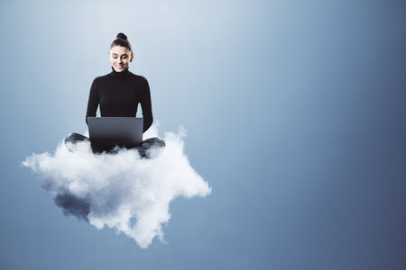 Woman sitting cross-legged on a fluffy cloud while using a laptop. Simple background. Concept of cloud computing, remote work, and technologyの写真素材
