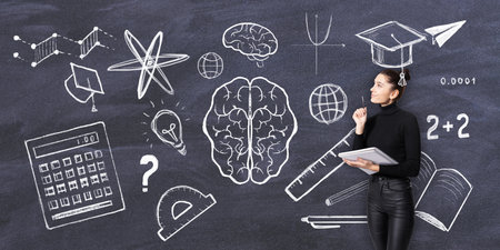 A woman holding a notebook stands next to a blackboard filled with chalk drawings of educational symbols and concepts, representing learning and knowledgeの写真素材