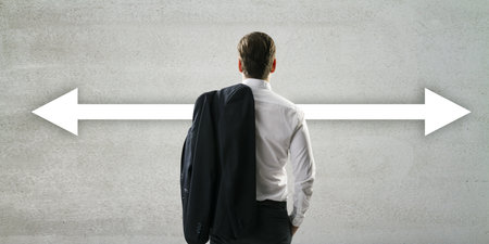 A businessman stands in a room with a concrete wall, where arrows point left and right, illustrating...