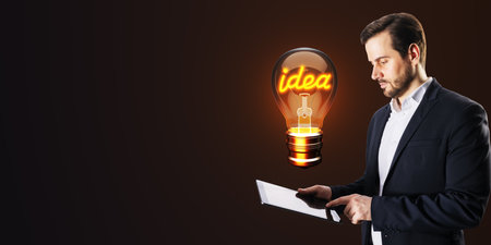 Businessman holding a tablet with a glowing lightbulb displaying the word idea on a dark background. Concept of creativity and innovationの写真素材