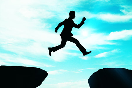 Silhouette of a businessman jumping between cliffs against a bright blue sky. Concept of ambition, goal achievement, and overcoming challengesの写真素材