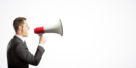Businessman in suit shouting into a megaphone on a clean white background. The image represents communication and announcement conceptsの写真素材