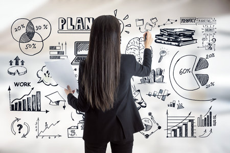 Businesswoman drawing business plan sketches on glass wall with hand-drawn charts and diagrams. Rear view. Blurred office background. Concept of planning and strategyの写真素材