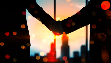 Silhouetted businesspeople shaking hands with a cityscape and sunset in the background, representing partnership and collaboration conceptsの写真素材