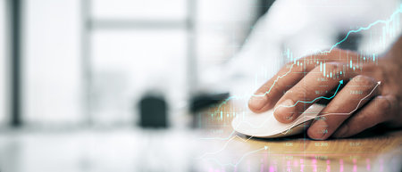 Businessperson's hand using a mouse on a wooden desk with financial graphs overlayed. Light...