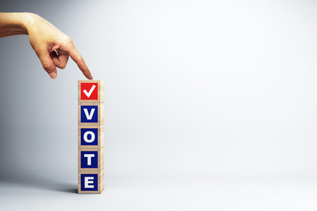 A hand points at a tower of wooden blocks spelling VOTE with a checkmark on the top block against a...