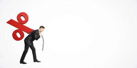 Businessman in a suit carrying a large red percentage symbol chained to his back, walking on a white background. Concept of financial burden, debtの写真素材