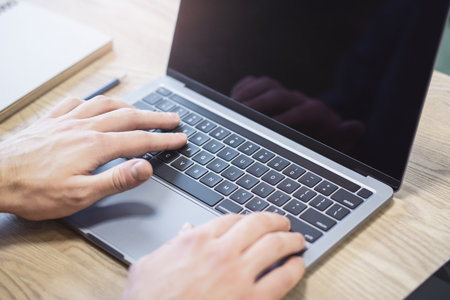 In a close-up shot, a man's hands are seen deftly typing on a modern laptop keyboard, set against a blurry office backdropの写真素材