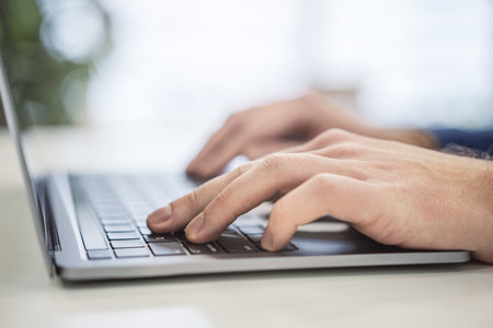 In a close-up, a man's hands can be seen typing on a modern laptop keyboard, with the office setting...